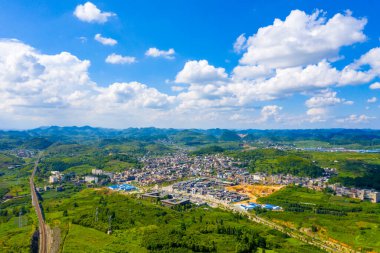 Guizhou Çin manzarası, beyaz bulutlu, açık mavi gökyüzünün altında yeşil tepelerin arasında yuva yapmış bir kasaba gösteriyor..