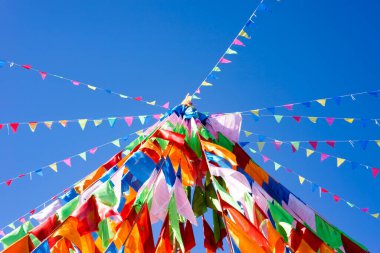 Parlak mavi gökyüzünde renkli bayraklar dalgalanıyor, neşeli bir atmosfer yaratıyor. Üçgen bayraklar birbirine bağlanmış, aşağıya doğru kıvrılan canlı bir gölgelik oluşturuyor, kırmızı, turuncu, sarı, yeşil, mavi gibi parlak renk spektrumlarını sergiliyor,