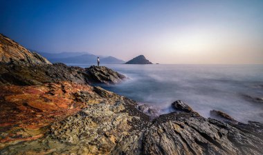 Yumuşak dalgalar kıyıya çarptığında okyanusa bakan engebeli kayalar üzerinde tek bir figür duruyor..