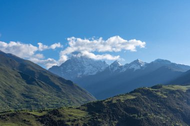 Karlı zirveler, açık mavi gökyüzünün altındaki yeşil vadilerin üzerinde görkemli bir şekilde yükselir..