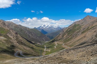 Çin Sichuan bölgesi dağ yolu yeşil vadileri geçerek mavi gökyüzünün altındaki karlı zirvelere doğru ilerliyor..