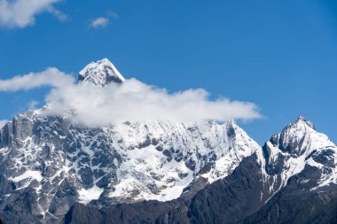 Çin 'in batı bölgesindeki mavi gökyüzünün altında karlı zirveler yükseliyor, bulutlar görkemli dağların etrafında sürükleniyor..