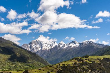 Karlı dağlar bulutlarla dolu açık mavi bir gökyüzüne karşı yükseliyor, Çin 'in Sichuan bölgesinin doğal güzelliğini gözler önüne seriyor..