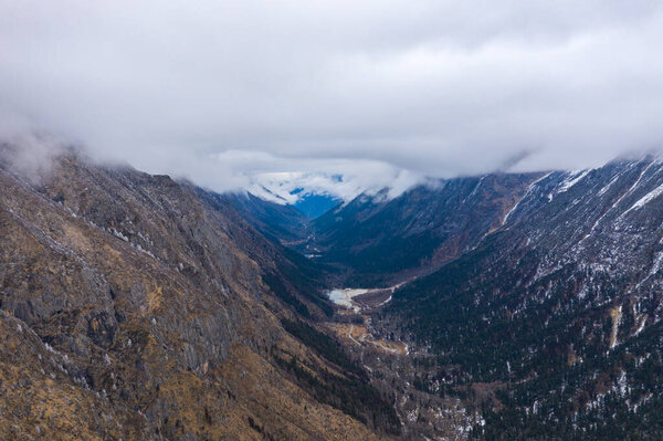 Огромная долина разворачивается под облачным небом, с прочными горными склонами, открывающими редкую растительность и участки снега, намекая на дикую красоту западных регионов Китая.