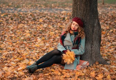 Güzel gözleri olan, şık gri bir ceket giyen ve parkta ağaçların yanında oturan kıvırcık, neşeli bir kız. Sonbahar zamanı. Portre.