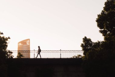 Seville, İspanya; 7 Temmuz 2024: Kadın gün batımında arka planda bir gökdelenle köprüden geçer. Arka planda Torre Sevilla olan Triana Köprüsü 'ndeki bir kadının silueti..