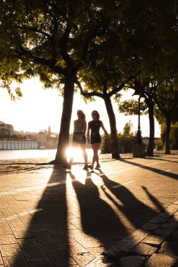 Seville, İspanya; 7 Temmuz 2024: Guadalquivir Nehri boyunca nehir kenarında gün batımında gezinen iki kadın. Gün batımında yürüyen iki kadının gölgesi.