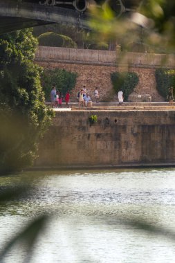 Seville, İspanya; 28 Ağustos 2024: Sevilla 'daki Guadalquivir Nehri' nin yanındaki gezinti alanında dinlenen insanlar.