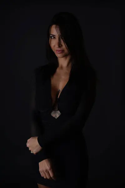 漂亮的年轻黑发女子穿着光滑的黑色连衣裙 在深灰色背景的衬托下 展示了迷人的领带 免版税图库照片