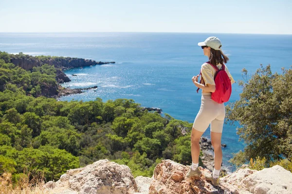 stock image The young woman with pink backpack is standing against mediterranean sea coast nature during the hike.