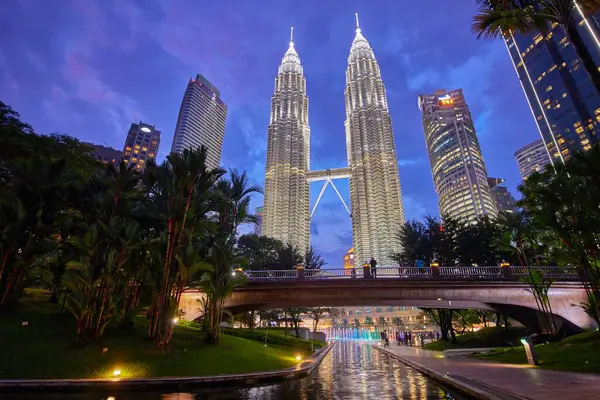 Kuala Lumpur, Malezya - 21 Haziran 2023. Petronas Kuleleri 'nin yanındaki tropikal şehir parkında geceleri palmiye ağaçları ve bitkiler olan gökdelenler.