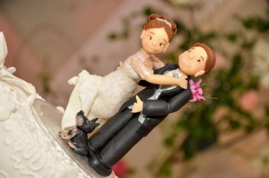 A charmingly detailed wedding cake topper featuring a bride and groom in elegant attire, with a playful black dog beside them, all set against a blurred green and pink floral background. clipart