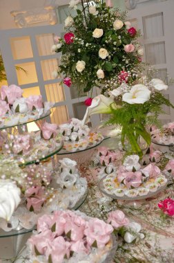 A beautifully arranged wedding dessert table adorned with an array of ornate sweets wrapped in pink, white and silver packaging. The backdrop features a lush floral arrangement of roses and lilies, adding elegance. clipart