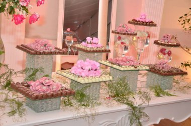 A sophisticated dessert presentation featuring an assortment of cakes adorned with pink floral decorations. The desserts are arranged on several glass stands with a reflective background. clipart