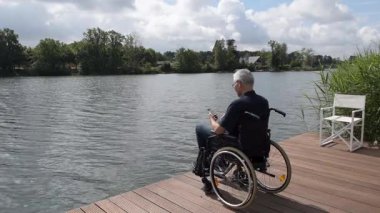 Wan 'daki yaşlı adam tekerlekli sandalyedeki yaşlı adam gölün kıyısındaki ahşap iskelede oturuyor. Akıllı telefonuyla fotoğraf çekiyor. Güneşli bir gün, su sakin ve karşı kıyıdaki yeşil ağaçlar...