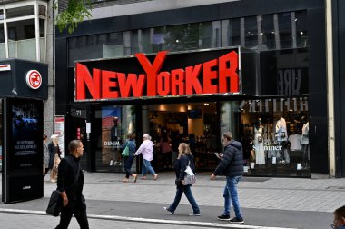 Düsseldorf, Almanya 21 Mayıs 2025 New Yorker. Bir giyim mağazasının önünden geçen yayaların olduğu şehir caddesi manzarası. Canlı kırmızı tabela dikkat çekiyor. Canlı bir alışveriş ortamı yaratıyor.