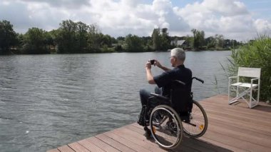 Wan 'daki yaşlı adam tekerlekli sandalyedeki yaşlı adam gölün kıyısındaki ahşap iskelede oturuyor. Akıllı telefonuyla fotoğraf çekiyor. Güneşli bir gün, su sakin ve karşı kıyıdaki yeşil ağaçlar...