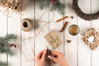 Kırpılmış kadın elleri, el yapımı karton kutular için halat kesiyor. Noel süslemeleri, hediyeler, köknar ağacı dalları ve çelenkler. Nakliye için mevcut kutuların doğal ambalajları ve ambalajları. Üst görünüm