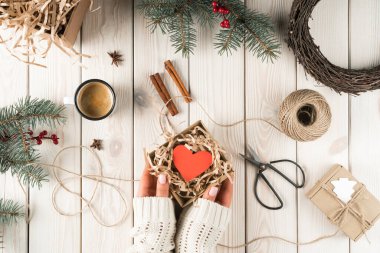 Kırpılmış kadın ellerinde beyaz kazakla elinde kırmızı kalple açık karton bir kutu tutuyor. Tarçın, makas, hediyeler, köknar ağacı dalları ve çelenk süslemeleri. Tatil için el yapımı hediyeler.