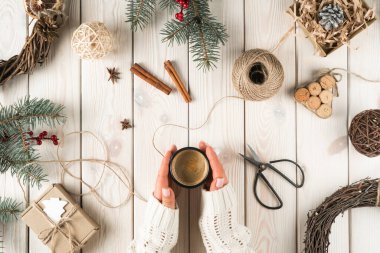 Tarçın, makas, köknar dalları, fındık ve çelenk gibi kostüm süslemelerinde sıcak bir fincan kahve tutan tanınmamış bir kadın. Evde Noel arifesi tasarımı ve el işi var. Üst görünüm