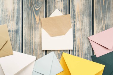 Gri ahşap arka planda açık kraft kağıt kartı olan beyaz bir zarf. Çok renkli zarflar yan yana uzanıyor. Uzayı kopyala Üst Manzara. Posta, yazışma, mesaj gönderme, tebrikler.