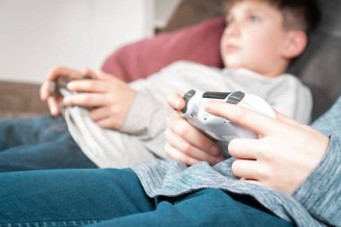 Çocuğun elleri modifiye edilmiş plastik kumanda kolu, seçici odaklanma. Çocuklar kanepede uzanmış video oyunları oynuyorlar. Kablosuz beyaz oyun konsolunu tutun. Gençlerin hobisi, yaşam tarzı. Modern teknoloji ve oyun.