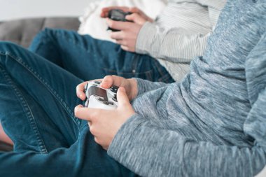 Oyun çubuklarıyla kanepede oturan adamların elleri, yakın görüş. Çocuklar video oyunları oynarlar. Kot ve uzun kollu erkekler evde dinleniyor. İlginç bir eğlence. En iyi arkadaşlar yaşam tarzı.