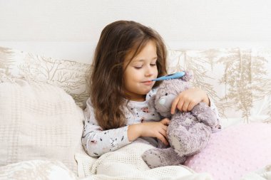 Gülümseyen, kendine güvenen, pozitif küçük çocuk, ağzında elektronik termometre tutan kız, oyuncak ayıyı kucaklayan ve yatakta sakinleştirici oyuncak. Covid 19, mevsimsel soğuk algınlığı, virüs enfeksiyonu. Boşluğu kopyala