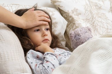 Yatak odasında tanınmayan bir kadının eli küçük kızın alnına dokunuyor. Üzgün, kayıtsız, sıkılmış küçük esmer kız beyaz yumuşak yatakta ayı oyuncağıyla yatıyor. Pediyatrist Sağlık ve Koruma