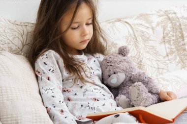 Tatlı, sevimli, pijamalı esmer kız yumuşak yatağında oyuncak ayıyla kitap okuyor. Evde öğrenme, eğitim ve bilgi. Hayal gücü ve gündüz rüyası. İçe dönükler için boş zaman etkinliği