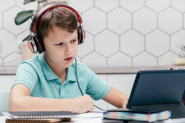 Tişört giyen, kulaklık takan, not defterlerinin yanındaki masada oturan, tablet ekranına bakan, online ders alan, dinleyen çocuk portresi. Uzaktan eğitim, uzaktan öğrenme.