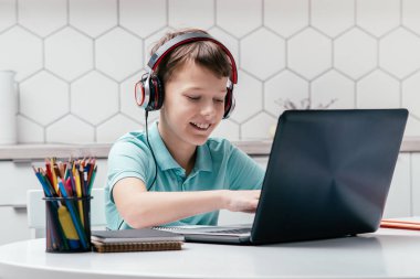 Tişört, kulaklık takan, kalemlerin yanındaki masada oturan, dizüstü bilgisayar kullanan, daktilo kullanan, oyun oynayan, mesajlaşan, müzik dinleyen çocuk portresi. Oyun, e-öğrenme, sosyal medya.