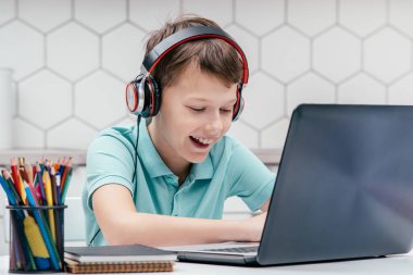 Tişört giyen, kulaklık takan, dizüstü bilgisayarın önünde oturan, daktilo eden, iletişim kuran, oyun oynayan, eğlenen çocuk portresi. Oyun, teknoloji, e-öğrenme, sosyal medya.