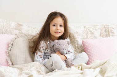 Uykulu gözlerle gülümseyen ve en sevdiği oyuncağı kucaklayan küçük kız. Uyanan bir çocuğun portresi. Çocuk sabah uyanır. Doldurulmuş oyuncaklarla uyuyorsun, oyuncak ayı. Rahat bir yatakta dinleniyor, uyanıyor..