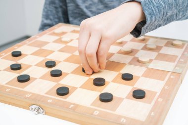 Ergenlik çağındaki bir çocuğun masada oturup tahta tahtada dama oynarken, planlama, inşaat stratejisi kurarken, elinde siyah bir dama taşırken çekilmiş bir fotoğrafı. Masa oyunları, geliştirme, eğitim, eğlence.