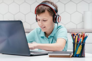 Tişört giyen, kulaklık takan, dizüstü bilgisayarın önünde oturan, sohbet eden, oyun oynayan, mesajlaşan genç bir çocuğun portresi. Oyun, e-öğrenme, sosyal medya.