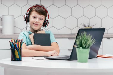 Mavi tişört giyen, siyah kırmızı kulaklık takan, dizüstü bilgisayarın önünde elinde not defterleriyle oturan genç bir çocuğun portresi. Uzaktan eğitim, teknoloji, e-öğrenim.