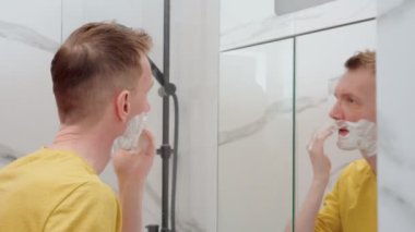 Yetişkin bir adam, banyoda yüzüne ve boynuna tıraş köpüğü sürer. Sakal tıraşı, sakal. Cilt rutini, dikkatli yüz yıkama. Gündelik giysiler içinde yakışıklı bir erkek aynaya bakıyor..