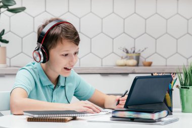 Kulaklık takan, not defterlerinin yanındaki masada oturan, tablet ekranına bakan, online ders alan, dinleyen, not alan, kalem tutan genç çocuğun portresi. Uzak öğrenim.