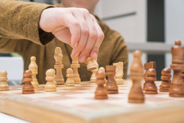 Çocuk yakın planda masa başında satranç oynuyor. Çocuğun eli tahta satranç tahtasındaki beyaz piyonu oynatıyor. Oyun stratejisi, taktikler. Ev eğlencesi, oyun zamanı, hobi. Eğitici ilginç tahta oyunu.