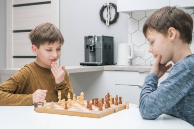 Okul çocuklarının mutfak masasında oturup satranç oynadığını düşünmek. Siyah ve beyaz figürlü tahta satranç tahtası. Oyun stratejisi, taktikler. Ev eğlencesi, oyun zamanı, hobi. Eğitim tahtası oyunu.