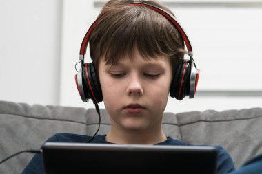 Tutkulu, dinlenmiş kulaklıklı esmer çocuk film izliyor, elinde dijital yastıkla kanepede oturmuş müzik dinliyor. Taşınabilir aygıt, müzik ekipmanı, ses kalitesi ve online oyun