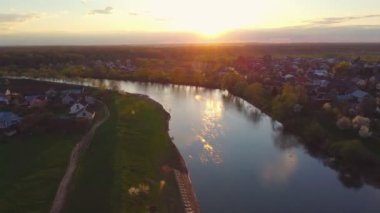 Nehir manzarası ve yaz akşamı hava manzaralı küçük bir kasaba. Kırsal yerleşim alanı ve kıvrımlı nehir kuşların gözünden görülebiliyor. İHA gökyüzünde uçuyor. Ufukta güzel gün batımı, cennet gibi şehir manzarası.