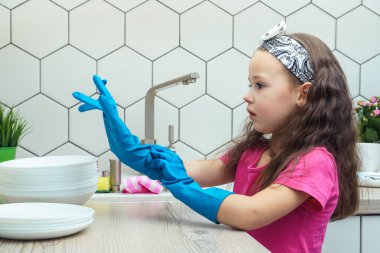 Ciddi, uzun siyah saçlı pembe tişört giyen, büyük mavi lastik eldivenler giyen, ellere bakan, mutfaktaki tabakları yıkamaya hazırlanan genç kızların yan görüntüsü. Ev işleri..