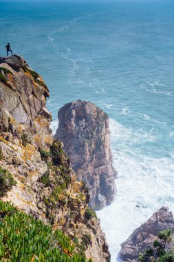 Küçük insan figürü yüksek uçurumun tepesinde duruyor, dalgalar aşağıdaki kayalara çarpıyor. Turizm ve seyahat. Eğlence ve eğlence. Yukarıdan bak. Deniz ve kayalar. Meditasyon ve rahatlama.