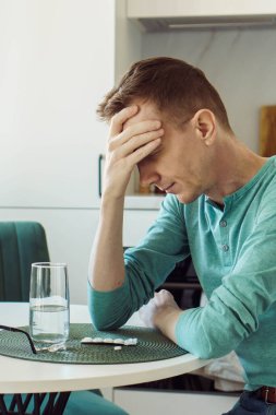 Baş ağrısı. Evdeki adam mutfak masasında oturuyor ve eliyle kafasını tutuyor, önünde bir bardak su var ve ağrı kesiciler, antidepresanlar var. Hastalık ve yalnızlık..