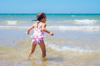 Beyaz mayo giyen, kumsalda suda oynayan, güneşli bir günde köpüklü deniz dalgalarıyla savaşan bronzlaşmış küçük bir kızın arka görüntüsü. Yaz, çocukluk, deniz, seyahat, tatil, tatil.