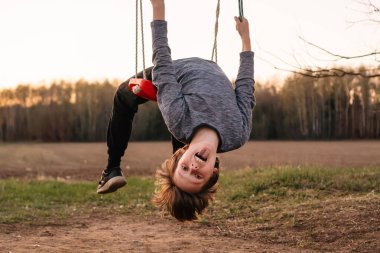 Hoş çocuk, gri uzun kollu, salıncaklarda oturan, ipi sıkıca tutan ve geriye doğru eğilen, kemer yaratan. Ergen kameraya bakıyor, dilini dışarı çıkarıyor ve eğleniyor..