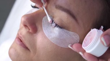 Kirpikçinin siyah kirpiklerini beyaz bantlarla kapağına pamuk tomurcuğu kullanarak pembe krem sürerken videosu. Kirpikleri dikkatlice yapıştıran bir kadın..