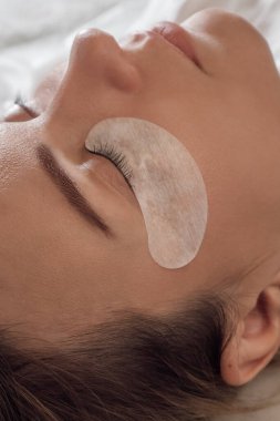 Kapalı gözlü ve koltukta uzanan göz kapaklarında beyaz yamalar olan muhteşem bayan müşterinin yüzünü kapatın. Siyah saçlı genç bir kadın kirpik uzatma prosedürüne hazırlanıyor. Dikey.